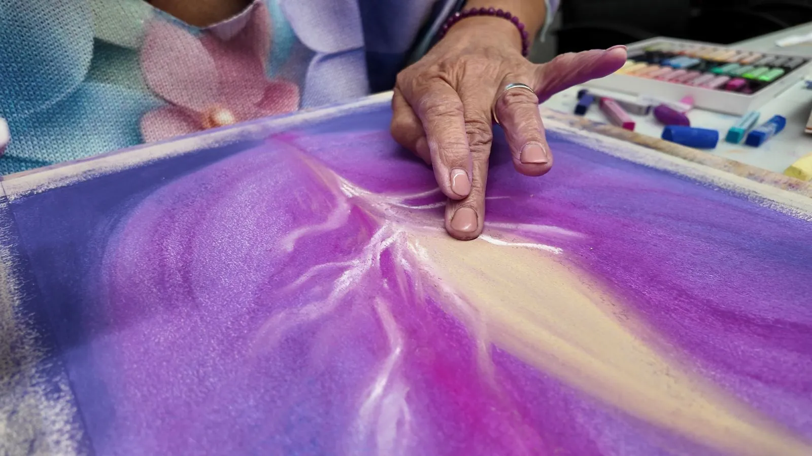 Une main travaille le pastel sec sur une grande feuille, à côté d'une boîte de pastels